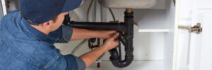 Professional plumber in blue cap and denim shirt installing or adjusting black P-trap pipe under bathroom sink.