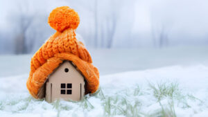 Small decorative house with orange knit hat and scarf sits in snowy landscape with frost-covered grass.