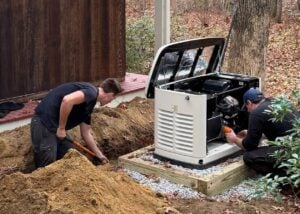 Technicians install a white Generac generator unit on a wooden platform next to a home foundation during outdoor service work.