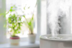 A white humidifier emits visible steam on a windowsill with potted plants and natural light in the background.