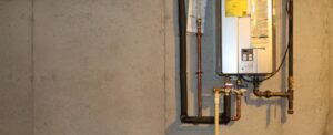 Tankless water heater unit mounted on concrete wall with copper piping, black inlet/outlet lines, and control panel with digital display.