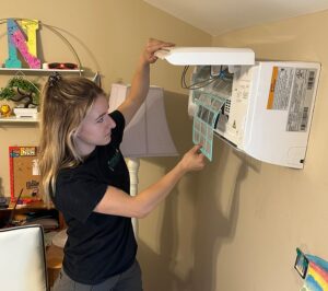 Woman replacing a green air filter in a white wall-mounted HVAC unit during home maintenance.