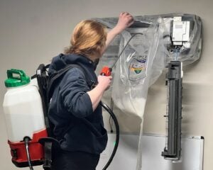 Technician performing air conditioning unit maintenance with spray equipment and tank on wall-mounted system.