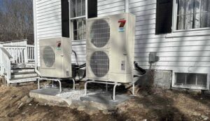 Two white Mitsubishi heat pump units installed on concrete pads against a house with white siding and black shutters.