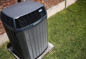 Gray air conditioning condenser unit mounted on concrete pad beside brick wall and green lawn.