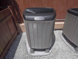 Gray and white air conditioning unit mounted on concrete pad beside brick wall and metal fence.