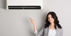 Woman in gray blazer smiling while adjusting wall-mounted air conditioning unit in home.