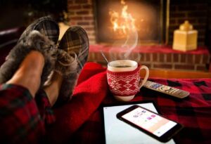 Person in cozy socks relaxing by fireplace with hot beverage in red knit mug, smartphone, and remote control on plaid blanket.