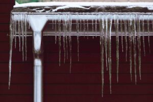 Frozen icicles drape from a metal gutter and downspout against a dark red building with snow-covered roof above.