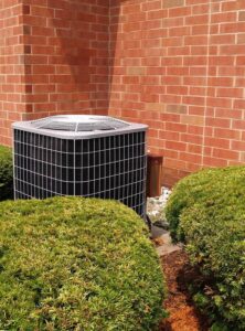 Black and white outdoor AC condenser unit mounted against red brick wall with green shrubs and gravel base.