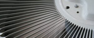 Close-up view of an air conditioner fan with metal fins and white housing showing intake vents and mechanical components.