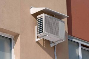 White outdoor air conditioning heat pump unit mounted on beige textured wall between two windows with protective overhang.