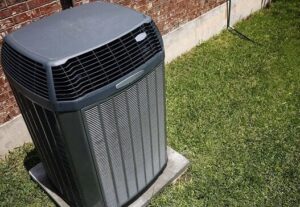 Gray central air conditioning unit installed on concrete pad beside brick wall and green lawn.