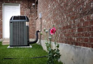 Gray HVAC air conditioning unit installed on grass next to brick house with pink flower blooming nearby.