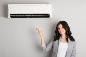 Woman in business casual attire holding a remote control while standing beneath a wall-mounted split air conditioning unit.