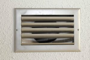White rectangular ceiling air vent with horizontal adjustable louvers and visible ductwork behind the grille mounted on textured wall.