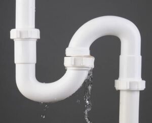 Close-up of white PVC S-trap plumbing pipe with water leaking from connection joint against gray background.