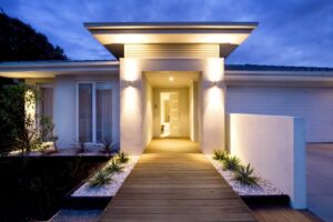 Contemporary home exterior featuring illuminated entrance, wooden walkway, potted plants, and garage at dusk.