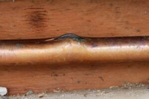 Frozen and cracked copper pipe with ice buildup and visible damage on wooden surface. Plumbing emergency.