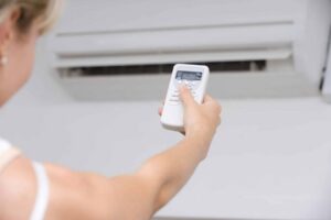 Person holding a white air conditioning remote control directed at a wall-mounted AC unit.