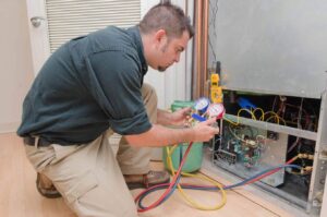 HVAC technician in blue shirt crouches while inspecting an air conditioning system with manifold gauges and colored refrigerant lines.