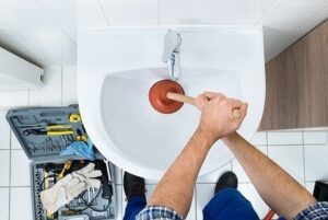 Professional plumber using a red plunger to clear a clogged bathroom sink drain while holding a toolbox.