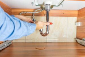 A plumber in a blue shirt repairs leaky pipes under a kitchen sink, adjusting connections on copper P-trap plumbing.