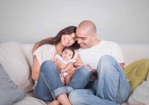 Parents and infant child smiling together on a white couch in a bright, comfortable home setting.