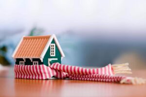 Small green house model with orange roof wrapped in a red and white striped scarf on a wooden surface, symbolizing home protection and warmth.