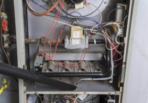 Interior view of HVAC furnace showing motor, electrical wiring, capacitors, and combustion chamber with technician performing maintenance.