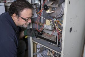 Technician wearing glasses and green gloves inspecting internal components of a residential furnace system with visible wiring and metal components.