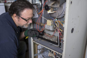 Experienced technician wearing glasses and green gloves inspects internal components of a residential furnace unit with visible wiring and mechanical parts.