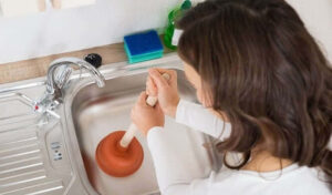 Woman using a red plunger to unclog a kitchen sink drain. Cleaning supplies and soap dispenser visible on the countertop.