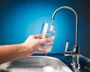 Hand holding a clear glass being filled with fresh water from a modern chrome kitchen faucet against a blue background.