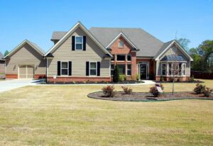 Large two-story residential home featuring tan siding, red brick accents, multiple gabled rooflines, and a two-car garage on manicured lawn.