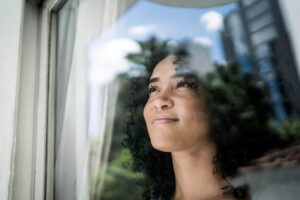 Woman gazing thoughtfully out a window with a blue sky and trees visible in the background.