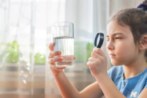 Young child in blue shirt holding a glass of clear water and examining it with a magnifying glass by a sunny window.