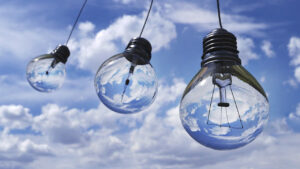Three hanging light bulbs with black bases reflecting a bright blue sky with white clouds against a cloudy background.