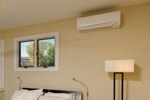 White ductless mini split air conditioning unit mounted on beige bedroom wall above bed with window and lamp nearby.