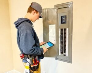 Technician in gray cap and blue hoodie holding tablet while inspecting residential electrical panel with circuit breakers and breaker switches.