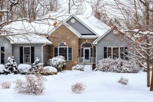 Modern residential house covered in snow with frosted evergreen shrubs, bare trees, and white pillared front entrance during winter.